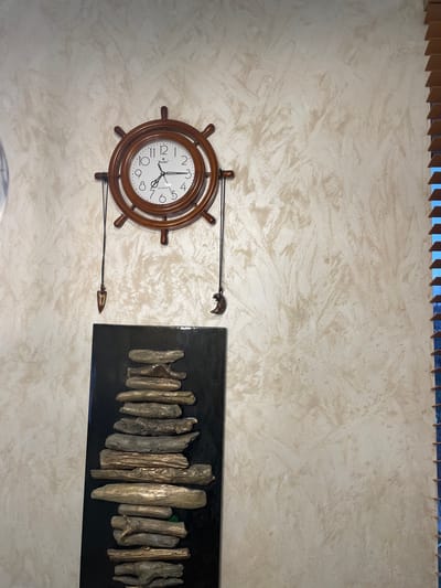 This image shows a wall with a decorative clock and an artwork in what appears to be an interior setting. The clock is designed to resemble a ship's wheel, with a brown wooden frame and black hands. The artwork to the right is a black rectangular piece featuring vertically arranged driftwood or similar objects of varying sizes, possibly adhered to the backing. The wall itself is textured, with a light beige or off-white color. There are no people visible in the image. The presence of Russian letters on the clock face possibly confirms it's location in Лесной Пейзаж Кп (шарапово), Russia.