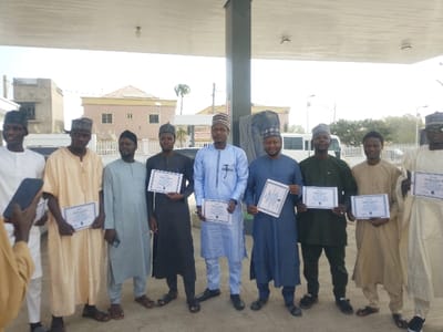Nine adult males are arranged in a horizontal line, facing the camera, under a white, open-air canopy structure in Kabuga, Nigeria. Each male is dressed in traditional Nigerian attire, consisting of long tunics and caps; garment colors include white, beige, light blue, dark blue, olive green, and black. All nine individuals are holding a white, framed certificate with dark borders at waist height, presenting them towards the foreground. Their facial expressions are generally neutral or exhibit faint smiles. In the lower left foreground, a hand holding a dark smartphone is partially visible. The background features multi-story buildings, including a prominent beige structure with multiple windows and a darker, unfinished concrete building. Partially visible vehicles, specifically a white van or bus, and streetlights are present further in the background under a bright sky.