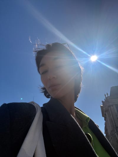 A portrait-oriented outdoor photograph features a woman's upper body and face against a clear blue sky. The sun is intensely bright in the upper right, generating significant lens flare that partially obscures the woman's face. She has short, dark, windswept hair and wears a dark grey coat over a lime green garment, with a white strap visible over her left shoulder. Her expression is neutral, and she looks directly towards the camera. In the extreme upper right background, a section of an ornate, dark stone building with classical architectural details and small statues along its roofline is discernible. This architectural style corresponds to the Palais de la Bourse, situated in the Place de la Bourse in Bordeaux, France. The environment is under strong natural daylight.