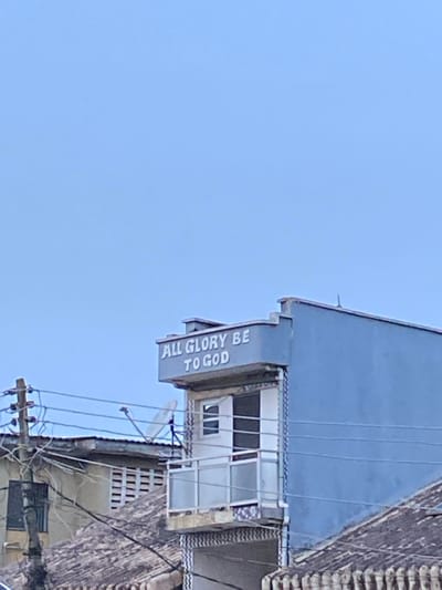 An image captured in Agege, Nigeria, depicts a pale blue/grey two-story building under a clear light blue sky. The building's upper facade displays the white capital letters "ALL GLORY BE TO GOD." A small, cantilevered balcony with a white railing is present on the second floor, featuring an open doorway and an adjacent smaller window. Below the balcony, the building's facade shows a vertical patterned section. To the left, a wooden utility pole with multiple black electrical wires extends upwards, with wires horizontally traversing the mid-ground. Partially obscured by these wires and the pole is a small white satellite dish. Corrugated reddish-brown roofs of adjacent structures are visible in the lower foreground and mid-ground on the left. No individuals are visible.