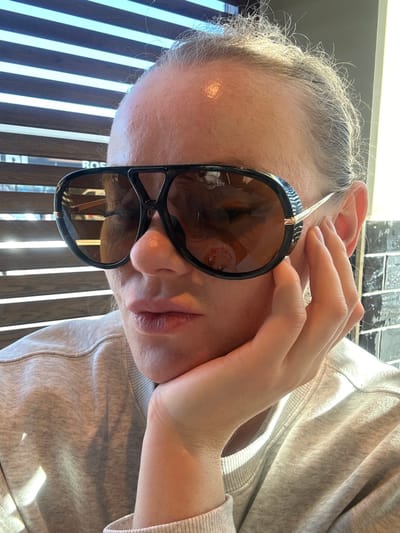A close-up, eye-level indoor photograph shows an individual, likely female, in Mysiadło, Poland. The person occupies the majority of the frame, positioned slightly to the left, with their head tilted slightly down and to the right. They are wearing large, dark-framed aviator-style sunglasses with brown-tinted lenses, obscuring their eyes. Reflections on the lenses indicate an indoor setting with horizontal window blinds or slats visible on the left side, and bright light entering. The individual's left hand supports their chin, with the palm resting against the jawline. They are dressed in a light grey crew-neck sweatshirt. The background to the left features horizontal light-colored slats or blinds with sunlight streaming through, creating distinct light patterns. To the right, a wall composed of dark, square tiles is visible. No other individuals are present or discernable.