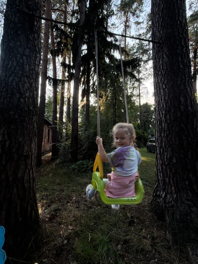 A young child is swinging on a bright green and yellow swing set in a wooded backyard. The child, who appears to be a toddler, is wearing a tie-dye t-shirt and a pink skirt. Their face is blurred due to motion, but they seem to be looking forward and enjoying the swing. The swing set is suspended between two large trees with rough bark. In the background, more trees and greenery are visible, along with a small brick building with a window. The sun is low in the sky, casting long shadows and creating a warm, late afternoon atmosphere. The overall scene suggests a playful and peaceful moment in a natural setting. The location is Udelnaya, Russia. No visible text.