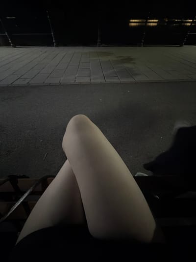This is a close-up, low-angle shot of a person's legs, which are covered in sheer pantyhose. The person is sitting on a dark surface, possibly asphalt or a rough pavement, which has some small white speckles on it. To the left of the person, there's a textured wall made of pale grey rectangular bricks laid in a horizontal pattern. The wall appears somewhat damp or stained in places. Beyond the brick wall, a dark metal structure, possibly a fence or gate, is visible. The light reflects off this structure, creating vertical yellow and white streaks, suggesting it might be nighttime or a very dimly lit area. The overall scene is dark, with the subject's legs being the brightest element, illuminated by an unseen light source. The lighting creates shadows and highlights on the pantyhose, emphasizing the form of the legs. The image composition focuses on the texture and light play on the fabric and the surrounding environment. No text is visible in the image.
