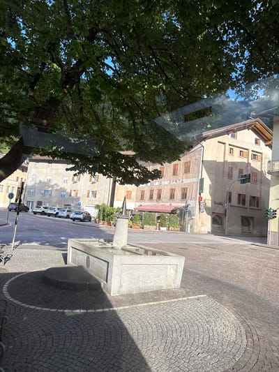 The image depicts a sunny street scene in Spormaggiore, Italy. In the foreground, a grey concrete fountain with an inscription that reads "2006 SPORMAGGIORE" is visible, situated on a cobblestone plaza. The plaza is shaded by a large tree with lush green leaves, its branches arching over the scene.
Several buildings line the street. To the left, a light-colored building with multiple windows and shutters is partially obscured by the tree. Further down the street, a taller, beige building with wooden shutters and a red and white striped awning over an outdoor seating area is prominent. This building appears to house a business, with text that may read "Bottega" and "Posta" visible on its facade. Adjacent to it, another beige building features a traffic light with green and red signals.
A row of parked cars, including a dark SUV, several white cars, and a silver sedan, are visible along the street. The street itself is paved with cobblestones, and a crosswalk is marked by white lines. The overall atmosphere is calm and serene, typical of a small Italian town.