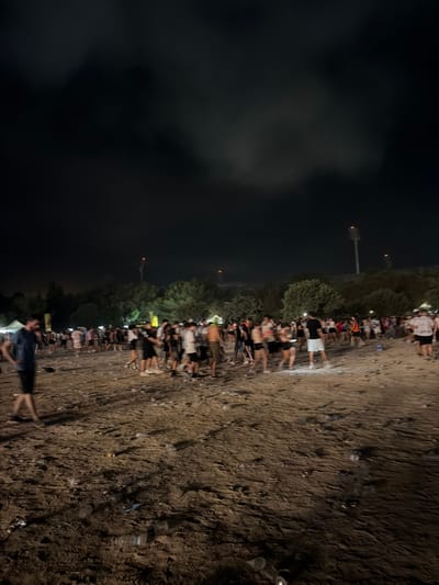 A nighttime photograph captures a crowd of people gathered on a sandy area littered with trash, primarily plastic bottles and cans. The scene appears to be outdoors, possibly at a festival or event, given the presence of tents and distant stadium lighting. The sky above is dark with some cloudy formations. The ground is uneven and sandy, with scattered debris throughout. Several individuals are visible in various states of motion, some standing in groups, others walking. The overall impression is of a post-event cleanup or a location where an event has recently concluded. The location is the Ta' Qali National Park in Mosta, Malta, a popular venue for outdoor concerts and festivals.