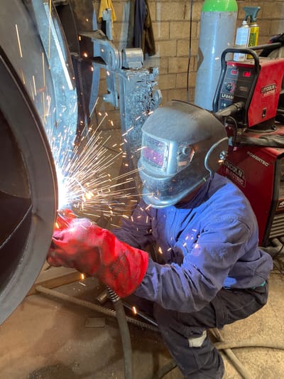 A person is welding a large, circular metal object in an indoor workspace. The welder is wearing a dark welding helmet, blue overalls, and red gloves. Sparks are flying from the point of welding. A red Lincoln Electric POWERTEC welding machine and a gas cylinder are visible in the background. The location is not specified as being in Zaljevo, Montenegro.