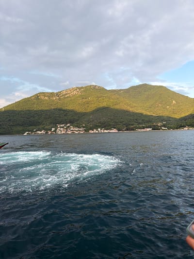 The image depicts a view of the Bay of Kotor near the village of Đurići, Montenegro. The foreground shows turbulent, foamy water, indicative of a boat's wake, with the deep blue water extending towards the shore. In the midground, a strip of coastline is visible, featuring a cluster of white buildings with red roofs, nestled against a lush green mountainside. The mountains dominate the background, with their slopes covered in dense vegetation and rocky outcrops. Sunlight casts a warm glow on the upper portions of the mountains, creating a contrast with the shadowed valleys. The sky above is partly cloudy, with patches of blue sky visible amongst the grey clouds. A hand holding a phone is partially visible in the lower right corner of the frame.