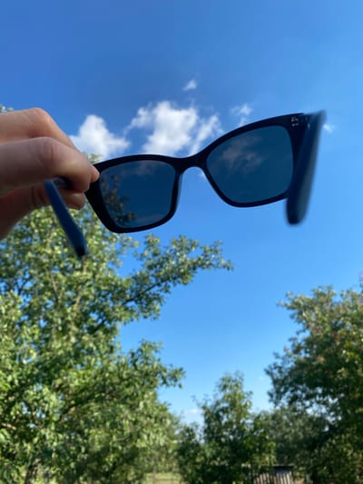 A hand holds a pair of black sunglasses up to a bright blue sky with scattered white clouds. The sunglasses have dark lenses that reflect the sky and trees. The hand is holding the arm of the sunglasses, which appears to be blue. Lush green trees frame the sky, with some branches and leaves in focus and others blurred. The sun appears to be shining brightly, suggesting a clear day. The scene is outdoors, with the sky and trees indicating a natural setting. The perspective is looking upwards, emphasizing the sky and the sunglasses. There is no visible text in the image. This photo could have been taken during daylight hours, possibly in the afternoon given the brightness.