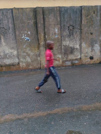 The scene is an outdoor setting in the city of Agani, Nigeria. A single young male individual, appearing to be of African descent, is captured in motion, walking from left to right across a dark asphalt surface. He wears a short-sleeved red shirt, dark blue athletic pants with two distinct light-colored stripes down the outer side of each leg, and black and white striped slide sandals. Pronounced motion blur is visible on his head and limbs, consistent with movement during capture.
The background features a textured, weathered concrete wall, segmented vertically into multiple panels. This wall exhibits discoloration, patches of apparent dirt or mold, and several torn paper fragments adhered to its surface. One panel of the concrete wall displays a circular relief carving resembling a multi-pointed star or gear. Above the concrete wall, a section of a plain, light brown or beige wall is visible. The asphalt ground appears damp in areas, and a horizontal streak of light brown sediment or discoloration runs across the lower mid-section of the frame. A small, reflective wet patch is present in the lower right foreground.