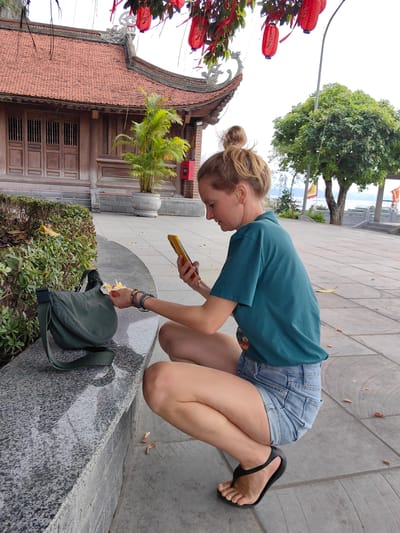 A woman is crouching on a paved area in Hong Gai Ward, Vietnam, with a building featuring a red-tiled roof and hanging red lanterns in the background. She is wearing a teal t-shirt, denim shorts, and black flip-flops. Her hair is pulled back in a bun. She is holding a yellow smartphone and appears to be taking a photograph of a white flower. A dark green messenger bag is resting on a stone ledge next to her. Foliage is behind the ledge. In the distance, a tree and flagpoles are visible.