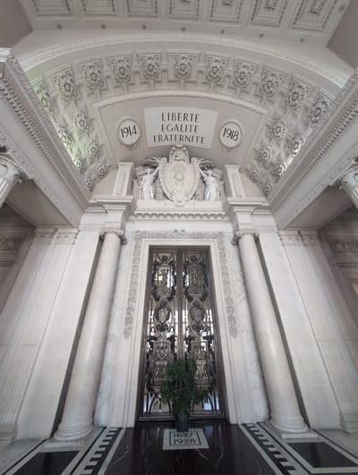 The image captures a wide-angle, low-angle view of an ornate, classical architectural interior. Dominating the scene is a grand, arched doorway with intricate metalwork, flanked by large, fluted marble columns. Above the doorway, a semi-circular inscription reads "LIBERTE EGALITE FRATERNITE" within a decorative frieze. Two circular motifs, one labeled "1914" and the other "1918," are positioned on either side of the inscription, possibly referencing historical events. Sculptural elements, including figures and crests, adorn the upper portions of the architecture, adding to the sense of grandeur. The ceiling is elaborately decorated with repeating floral and geometric patterns. The floor is a polished dark marble with a contrasting black and white patterned border. A potted plant with large green leaves is placed to the left of the doorway, and a small white plaque on the floor near the plant displays the numbers "1927" and "1928". The overall impression is one of solemnity and historical significance, indicative of a public building or monument. The location is identified as the war memorial in Urvillers, France.