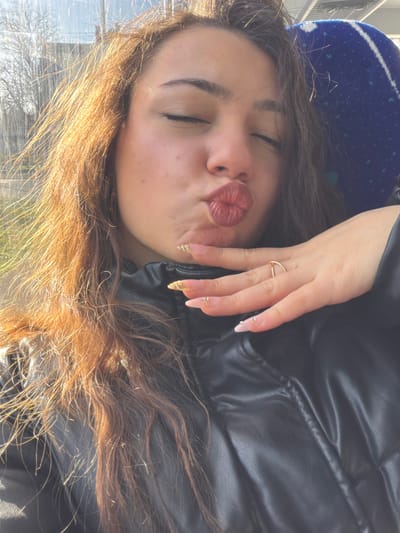 A close-up selfie depicts a female individual inside a vehicle, located in Bouxwiller, France. The subject, positioned centrally, has reddish-brown, wavy hair illuminated by strong sunlight from the left, creating a luminous effect on the strands. Her eyes are closed, and her lips are puckered into a prominent "kiss" pose. She wears a black, possibly faux leather, puffer jacket with visible stitching across the chest. Her left hand is raised, with fingers curled and thumb extended upwards, positioned near her chin. A thin, gold-colored ring adorns her left ring finger. Her almond-shaped fingernails exhibit a clear or pale pink base with white tips, gold thread-like accents on the index and middle fingers, and small rhinestones near the tips of the ring and pinky fingers. In the background, to the right of her head, a blue seat with a distinct patterned fabric is visible, suggesting a bus or train interior. Through a window on the left, bare tree branches and a distant light-colored structure are faintly discernible against a bright sky, indicating an outdoor environment under daylight conditions.