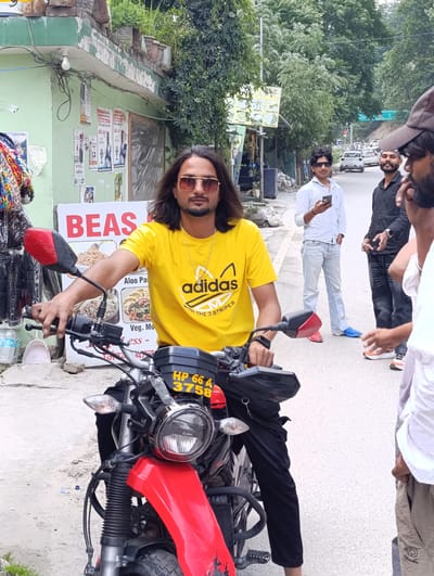 A man with long, dark hair and sunglasses is sitting on a red motorcycle. He is wearing a yellow t-shirt with an Adidas logo and black pants. The motorcycle has the license plate "HP 66 A 3758". Behind him, a sign reads "BEAS" and lists food items such as "SIDDU", "MOMO'S", "BREAD OMELETE", "Aloo Pa", "ICE", "MAGGI", and "DAL CHAWAL".
In the background, other people are visible, including a man in a white shirt holding a smartphone and another man in a black shirt. The scene appears to be a street in Manali, India, with trees lining the road and a slightly overcast sky. There are shops and buildings along the street, with posters and advertisements on the walls. The overall impression is a casual street scene, possibly a tourist area.