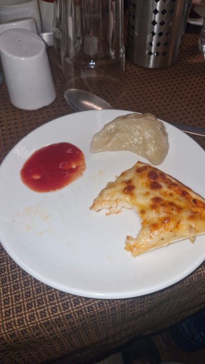 A white plate rests on a brown patterned tablecloth. On the plate are three food items: a slice of pizza with melted cheese and a bite taken out of it, a single steamed dumpling, and a small pool of red sauce, likely ketchup. In the background, out of focus, are elements of a dining setting, including a clear glass, a stainless steel utensil holder, and a white ceramic container. The overall scene depicts a casual meal. The image does not contain any specific visual cues to indicate its location in Manali, India.