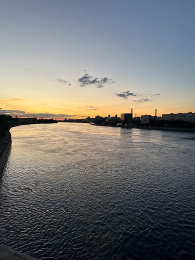 This is a horizontal, eye-level photograph capturing a wide river at sunset. The sky transitions from a pale blue at the top to a vibrant orange and yellow near the horizon, with a few scattered clouds. On the left bank of the river, a curved embankment with a walkway and trees is visible, leading towards buildings in the distance. The right bank features a cityscape with various buildings, including some with smokestacks, and streetlights. The river's surface is dark and reflects the colors of the sky, with visible ripples and waves. The overall impression is a peaceful urban scene at dusk. While the prompt mentions the city of Romanovka, Russia, there are no specific visual cues in the image that definitively confirm this location.