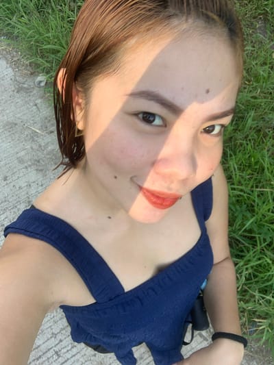 The media file features a close-up, top-down shot of a young woman lying on her back outdoors. She has light brown hair, styled to fall around her face, and her head is tilted to her left. Her eyes are open and looking towards the camera, and she is wearing coral-colored lipstick. A shadow, possibly from sunlight filtering through leaves, casts a diagonal line across her face from her forehead down to her cheek. Her skin appears fair with a scattering of freckles on her cheeks and a mole on her right cheek. She is wearing a dark blue, sleeveless top with textured fabric. Her right arm is bent, with her hand resting near her head, and she wears a black elastic band on her wrist. Her left arm is also bent, and the image crops to show her upper chest and shoulder. The background consists of green grass and a concrete surface, suggesting an outdoor setting. The lighting indicates it is likely daytime, possibly late afternoon given the warm tones of the shadow. The overall mood appears relaxed and casual, possibly a selfie taken while enjoying a quiet moment outdoors in Kiblawan, Philippines.