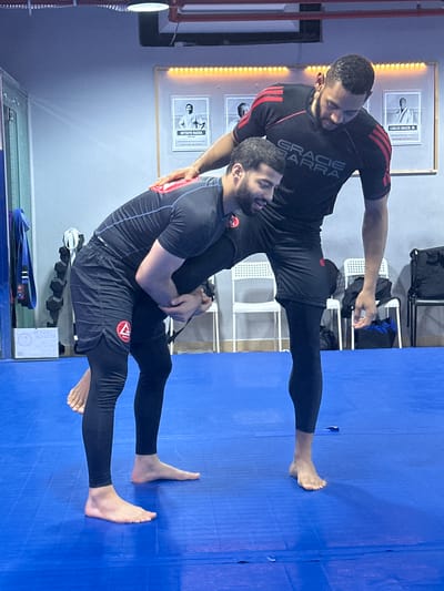 Two men are engaged in a martial arts grappling technique, likely Jiu-Jitsu, on a blue mat indoors. The man standing is darker-skinned, wearing a black shirt with red stripes and the words "Gracie Barra" visible. The man being lifted wears a dark top and shorts over leggings. The location is most likely a Jiu-Jitsu studio in دبي, United Arab Emirates. Background details include framed posters on the wall and chairs.