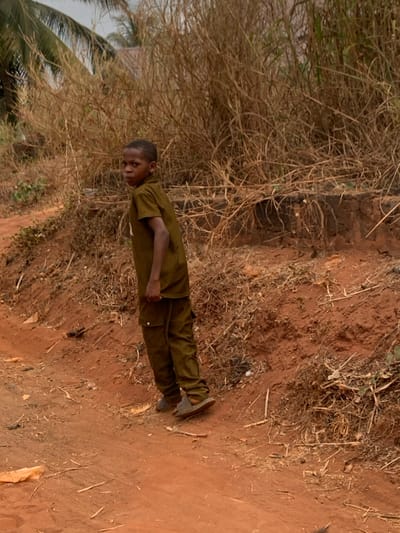 A male child, estimated to be between 8-12 years old, stands on a reddish-brown dirt path in Aguleri, Nigeria. He is wearing a dark olive-green short-sleeved collared shirt and matching cargo-style pants, with light-colored slide sandals. The child is oriented with his body angled slightly away from the camera, but his head and upper torso are turned, and he is looking directly towards the camera. His left arm is bent at the elbow, with the hand partially clenched. The ground consists of reddish-brown soil, mixed with dry leaves, twigs, and minimal debris. Behind and to the right of the child, an elevated bank of reddish soil supports dense, dry, straw-colored vegetation. A dark, low-lying structural element, possibly concrete or hardened earth, is visible at the base of this bank. In the upper left background, green palm fronds are visible against an overcast sky.
