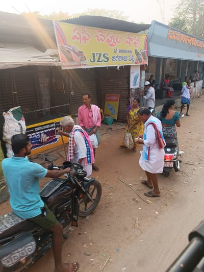 A street scene in Kothacheruvu, India. A man on a motorcycle is in the foreground. In the midground, an elderly man with a walking stick walks past a yellow tobacco advertisement toward other people. One woman stands near a scooter. Further away, people stand in a shaded area. Above are signs in Telugu and advertising for shoes. The area appears to be a roadside stop.
