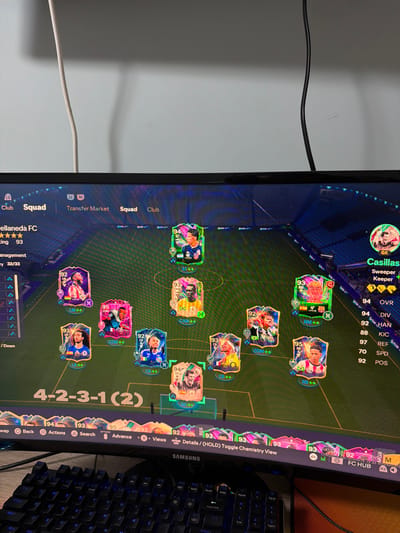 This is a close-up, eye-level, indoor shot of a computer monitor displaying a video game screen, likely EA Sports FC 24, given the interface. The game is in squad selection mode, showing a soccer team formation labeled "4-2-3-1 (2)". The screen displays player cards with their ratings and positions, including a goalkeeper labeled "Casillas" with a rating of 94. The team name displayed is "Gelllaneda FC". The monitor is positioned above a black Samsung keyboard with blue backlighting, which is partially visible in the foreground. The overall environment appears to be a room, with a white wall and a black cable visible in the background. There is no indication of the city of Pavolche, Bulgaria, or any specific geographic location within it.