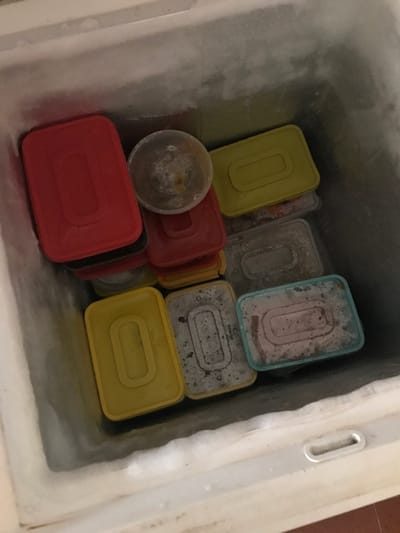 The image displays the interior of a chest freezer, viewed from directly above. The freezer's internal walls are heavily coated with a thick layer of white frost. The space is densely occupied by various plastic food storage containers, all appearing frozen.
Identified containers include:
- Multiple rectangular containers with opaque red lids.
- Multiple rectangular containers with opaque yellow lids.
- Several rectangular containers made of clear or translucent plastic, many showing significant frost accumulation on their surfaces. One of these translucent containers features a light blue rim.
- A single small, round, clear plastic container, also heavily frosted, positioned near the upper-center of the arrangement.
The contents of the containers are largely obscured by their lids or layers of frost, although some appear to contain dark or reddish food items. No individuals are present in the frame. The scene is located in Calabar, Nigeria.