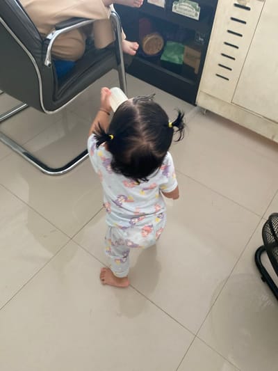 A toddler with dark hair in pigtails wearing a light-colored pajama set with cartoon characters is holding a white plastic cup and standing on a beige tiled floor. The toddler is facing away from the camera and is in focus. To the left, a part of a black office chair with chrome legs is visible, with a person's legs clad in beige trousers and a blue cushion underneath. In the background, a beige cabinet with a dark section containing shelves of items is present. To the right, the edge of a black fan is visible. The lighting appears to be artificial, creating reflections on the tiled floor. The overall scene depicts a domestic indoor setting.