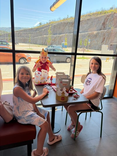 Three children are seated at a table in a Burger King restaurant. The child on the left, a girl with long brown hair, wears a light blue and white tie-dye dress and light pink slides. She is looking towards the camera and smiling. The child in the middle, a boy wearing a golden Burger King crown and a red and yellow shirt, is making a rock-on hand gesture and smiling. He is holding two Burger King Jr. meals. The child on the right, a girl with long brown hair, wears a white t-shirt with a graphic design and black shorts with white trim. She is looking towards the camera and smiling. On the table, there are two Burger King Jr. meals, a small red tray with food, and a pink bag. The restaurant has large windows with a view of a rocky hillside and cars parked outside. The floor is tiled, and the seating appears to be upholstered in dark red. The overall scene suggests a casual meal outing.