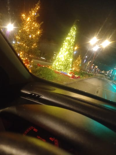 Captured from inside a vehicle, the image shows a night scene. Bright lights illuminate Christmas trees adorned with yellow and green lights, visible outside. The car's dashboard, including the steering wheel and speedometer, dominates the foreground, the speedometer display in red. The image is slightly tilted, creating a dynamic perspective.