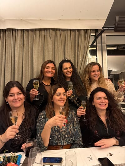 Six smiling women are gathered at a table. Some hold champagne flutes and pose for a photo. A "Fabulous" sash is visible among them. The table is covered with a white tablecloth and scattered with confetti. A mobile phone rests on the table, displaying a time of 23:17. Flowers in a vase are situated at the table's edge. A beige curtain is visible in the background.