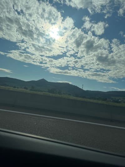 The image captures a daytime landscape viewed from the interior of a moving vehicle. The upper two-thirds of the frame is dominated by a bright blue sky, partially obscured by extensive white and light grey stratocumulus clouds. The sun is visible in the upper right-center, appearing as a brilliant, diffuse light source partially veiled by clouds.
A range of dark green to grey hills forms the horizon line across the middle of the image. On a prominent peak in the central-left section of these hills, a distinct stone structure, identified as a castle (consistent with Château de Kintzheim), is silhouetted against the sky. Further to the right, a tall, red-brown lattice transmission tower stands on another hill.
In the immediate foreground, a grey concrete highway barrier runs horizontally. Beyond this barrier, a narrow strip of green grass with sparse trees and shrubs precedes the base of the hills. The bottom-most portion of the image shows a dark asphalt road surface with a single white solid line visible on its left edge. The extreme bottom edge of the frame reveals the dark, curved interior trim of a vehicle's window or door. No people, vehicles other than the perspective vehicle, or animals are visible. The scene is consistent with the area around Kintzheim, France, given the identifiable castle.