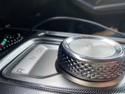 The image shows a close-up view of a car's gear selector. The selector is a rotary dial with textured sides and a metallic finish. The dial is situated in a car's center console, which is made of black material with a carbon fiber pattern. The gear selector indicators, "R", "N", and "D/M", are visible on the console. In the background, the interior of the car is visible, including the dashboard and possibly a display screen. The lighting suggests it is daytime, and the car is parked indoors or in shade. There are no people or animals visible in the image. The exact location in the city of Surgut, Russia, is not discernible from the image.
