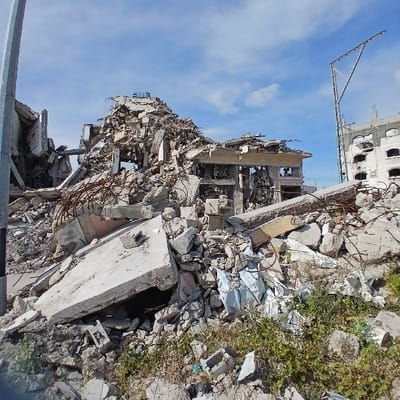 This image presents a scene of extensive structural destruction in Jabalia, Palestinian Territory. The foreground and midground are dominated by a large mound of rubble, consisting primarily of fractured concrete slabs, broken blocks, and twisted steel rebar. This debris appears to be the remnants of collapsed multi-story buildings. In the center-midground, a partial structure with exposed internal elements and empty window or door frames remains somewhat intact amidst the surrounding wreckage. To the far left, a grey utility pole stands vertically. To the right, a partially damaged, multi-story concrete building with numerous dark, empty window openings is visible in the background, alongside a tall, skeletal metal framework that resembles a structural support or pole. The ground in the immediate foreground shows sparse patches of green vegetation growing through the rubble. The sky above is clear blue with minimal scattered white clouds. No individuals are present in the frame.