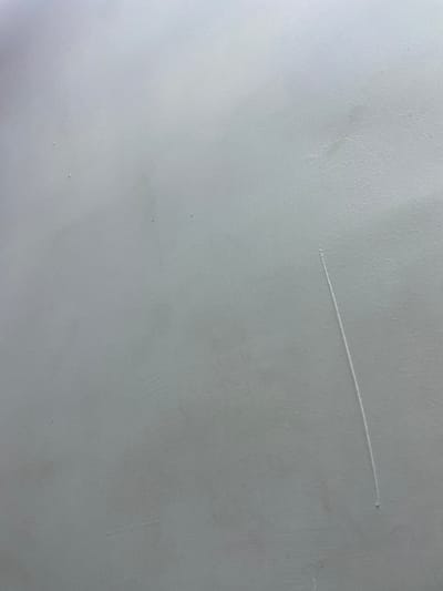 This is a close-up, top-down photograph of a textured, pale gray surface, possibly a wall or ceiling. A single, thin, diagonal scratch is visible, running from the upper left to the lower right of the frame. The scratch appears to be a superficial mark on the surface, with no indication of damage to a substrate. The lighting suggests an indoor setting, with the light source likely originating from above and to the left, casting subtle shadows that highlight the texture of the surface. There are no people, objects, or discernible location cues within the frame. The overall impression is one of a neutral, unremarkable surface. No text is visible in the image.