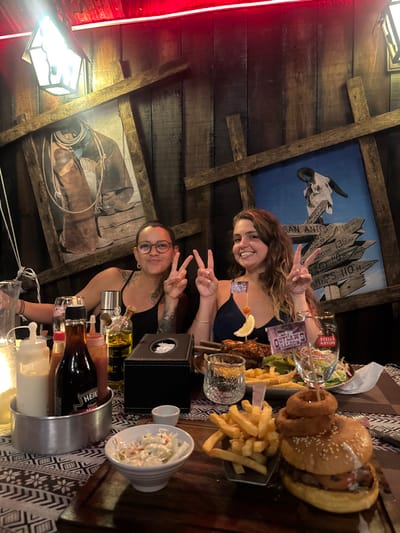 Two adult females are seated at a table in Outlaw's Saloon & Steakhouse, located in Moo 1, Rawai, Mueang Phuket District, Phuket 83130, Thailand. Both individuals are smiling and holding up peace signs with both hands towards the camera.

The female on the left has short, dark hair, wears glasses, a black tank top, and visible tattoos on her left arm. The female on the right has long, wavy brown hair and wears a dark blue spaghetti strap top.

The restaurant's interior features a dark wooden paneled wall. Two large, framed pictures are mounted on the wall; the left picture depicts a close-up of cowboy boots and chaps with a coiled rope, while the right picture displays a bull skull against a blue background, surrounded by directional signs pointing to various Western American locations like "SAN ANTONIO 139 mi" and "ROUTE 66 39 mi." A red neon strip light runs along the top of the wall, and a rustic lantern-style light fixture is visible on the upper left.

The table is set with a patterned placemat. On the table, a silver caddy contains condiment bottles, including Heinz sauce and two squeeze bottles with white and red contents. A clear glass bottle of yellowish liquid (possibly oil or dressing), a black tissue box, and a small white bowl of coleslaw are also present. A large platter in front of the right female holds glazed ribs, a generous portion of french fries, and a side salad. The ribs are garnished with a lemon slice and a cherry tomato on a toothpick. A small flag labeled "OUTLAW'S SALOON" is stuck into the ribs, and another identical flag is in front of the right female. An empty Stella Artois branded glass and another patterned glass are also on the table. In the foreground, a wooden serving board holds a large burger topped with onion rings and an additional side of french fries.