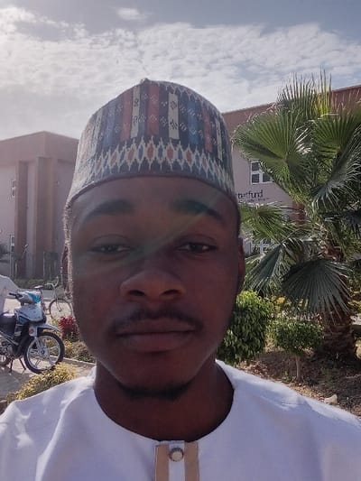 A young adult male, of apparent African descent, is depicted in a close-up, outdoor photograph taken in Kwazailewa, Nigeria. He wears a white, collared garment with a decorative tan and white buttoned placket and a patterned cap (kufi-style) featuring horizontal bands of red, blue, white, and geometric motifs. He has a moustache and a chin beard, with a neutral facial expression. The sky above is bright with scattered white clouds. In the background, to the subject's left, a light-colored building is visible alongside a parked white and blue scooter and a partially obscured bicycle. A second individual in white attire is partially visible to the far left. To the subject's right, another light-colored building displays partially visible text including "tetfund," accompanied by a large palm tree with fan-shaped fronds and various green shrubs.