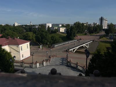 The image captures a cityscape in Vitsebsk, Belarus, likely from an elevated viewpoint looking towards a river and a bridge. In the foreground, a grand stone staircase with ornate railings and spherical finials leads downwards. Two black lampposts with multiple globes stand sentinel along the staircase. To the left, a light yellow building with a red roof and white trim is visible, partially obscured by dark green evergreen trees.
Beyond the staircase, a paved plaza stretches out, dotted with pedestrians. A white pedestrian bridge with arched supports spans a body of water, connecting the plaza to an area with more greenery and buildings. Further in the distance, the city skyline is characterized by a mix of architectural styles, including modern high-rise buildings, institutional structures with green roofs, and a prominent tall white building with a darker facade and a sign at its apex. A clock tower with a pointed spire is also visible amidst the trees in the mid-ground. The sky is a clear blue with a few wispy clouds. The scene depicts a public urban space with a blend of historical and contemporary elements, suggesting a pleasant day with moderate pedestrian activity.