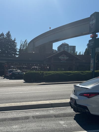 A white Acura sedan is parked on a city street in Burnaby, Canada. The car is facing away from the camera and is partially obscured by a concrete barrier. In the background, a White Spot restaurant is visible, with a sign that reads "White Spot Since 1928". The restaurant has a rustic, log-cabin-style facade and an outdoor patio with tables and umbrellas. An elevated highway overpass runs above the restaurant and the street. The sky is clear and blue. The scene appears to be set during the daytime.