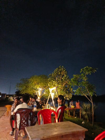 The image captures an outdoor seating area at night, likely a restaurant or cafe, in Purbachal, Bangladesh. The scene is illuminated by strings of warm-toned lights draped across trees and bamboo structures, creating a cozy and inviting ambiance. Several groups of people are visible, mostly young men, engaged in conversation and enjoying their time.
In the foreground, a wooden table is set up with red chairs around it, suggesting a dining or gathering space. One person is seated facing away from the camera, looking towards the table. Another red chair is prominently featured on the right. Further back, a group of men are seated at tables, with some appearing to be in conversation. One man is leaning forward, seemingly engaged in discussion, while another is looking away. A bottle of water is visible on a table in the middle ground.
The background reveals a dark sky, indicating it is nighttime. The presence of trees with their leaves lit up by the decorative lights adds to the aesthetic appeal. A body of water is visible to the right of the frame, suggesting the location might be near a lake or river. In the distance, some structures and lights hint at a more urban or developed area. The overall mood appears relaxed and social, with people enjoying a casual evening outdoors. The weather seems clear, as indicated by the visibility of the night sky.