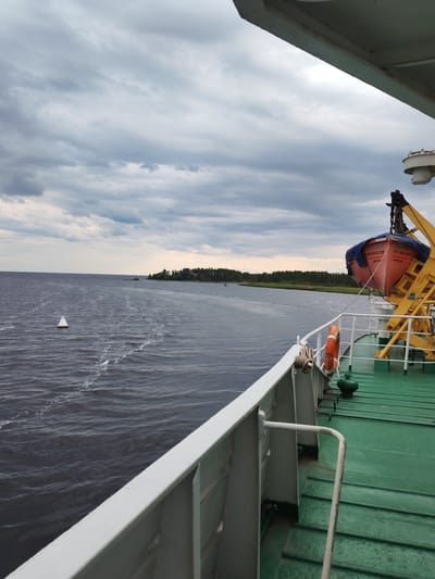 The image shows a view from the deck of a ship in the city of Shal'skii, Russia. The sky is overcast with dark clouds. The sea is choppy with whitecaps. A small white buoy is visible in the water. In the distance, there is a treeline along the shore. On the deck of the ship, there is a lifeboat hanging from a davit. A life preserver is also visible on the deck. The deck itself is painted green. The railing of the ship is white.