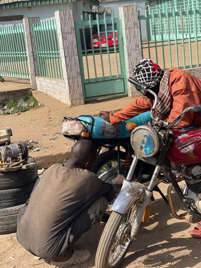 In Chikaji, Nigeria, an outdoor scene depicts two adult males engaged in motorcycle repair on a dirt ground. The individual on the left, wearing a dark brown tunic and trousers with light-colored clogs, is crouched, facing away from the camera, attending to the rear wheel area of a light blue motorcycle. Adjacent to him on the left, a stack of three tires supports a metallic engine component. The second individual, positioned on the right, wears an orange long-sleeved shirt, a plaid neck wrap, and a black and white mesh-patterned beanie. He leans over a red "Boxer" brand motorcycle, its front headlight adorned with a blue sticker, focusing on its front section. Red sandals are visible near his feet. The background features a light green metal fence and gate with tiled pillars. Through the open gate, a red vehicle is visible. Scattered refuse is present on the dirt surface to the far left. The scene is brightly lit by natural sunlight, casting shadows.