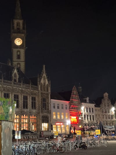This image captures a nighttime view of the Korenmarkt in Ghent, Belgium. The iconic belfry tower, illuminated by a warm light emanating from its clock face, stands prominently on the left. Adjacent to it, a row of historic buildings with ornate facades and arched windows line the square. Many of these buildings are lit from within, casting a red glow onto the street.
A Burger King restaurant is visible with its distinctive red signage. In the foreground, a large cluster of bicycles is parked at racks, suggesting a popular mode of transport in the city. Several outdoor seating areas for cafes and restaurants are set up, some with umbrellas, indicating a lively evening atmosphere. A few people are present, appearing to be walking or seated, contributing to the urban ambiance. The sky is dark, indicating it is nighttime.