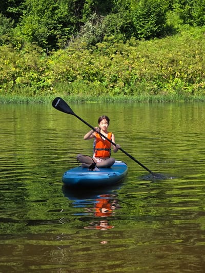 A young girl sits on a blue paddleboard in the middle of a calm, green body of water, holding a paddle. She is wearing an orange life vest and appears to be paddling. The paddleboard is in focus, as is the girl. In the background, a lush green forest lines the edge of the water. The scene is brightly lit by the sun, casting reflections on the water's surface. The location is the Voinskaya Chast' Mchs, Russia.