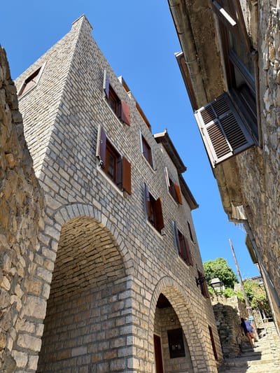 A low-angle shot captures a narrow street in the old town of Ulcinj, Montenegro, with two stone buildings flanking the pathway. The building on the left, constructed of light-colored stone bricks, features arched entrances at its base and several windows with dark wooden shutters, some of which are open. The building on the right, also made of stone, is positioned at an angle, with a portion of its facade and a sloped roof visible. A person in a blue shirt and dark shorts is walking up a stone staircase that ascends between the two buildings, with a bicycle leaning against a wall nearby. Lush green trees and more stone structures are visible in the background under a clear blue sky.