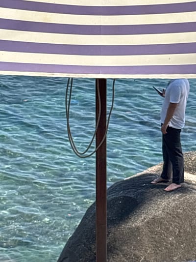 The image depicts a person standing on a rock formation next to calm blue water, likely the sea, in Kamala, Thailand. The person wears a white shirt, black pants, and possibly flip-flops, and appears to be using a phone. A metal pole is situated horizontally in the foreground, with hanging wires near it. A purple and white striped fabric covers the left side of the frame, potentially part of a beach umbrella or similar structure.