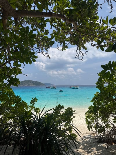 The image depicts a beach scene in Lam Kaen, Thailand. The view is framed by foliage in the foreground. Turquoise water stretches toward the horizon where clouds partially obscure the sky. Several boats are visible in the water. A sandy beach is present to the right of the water. An island can be seen in the distance.