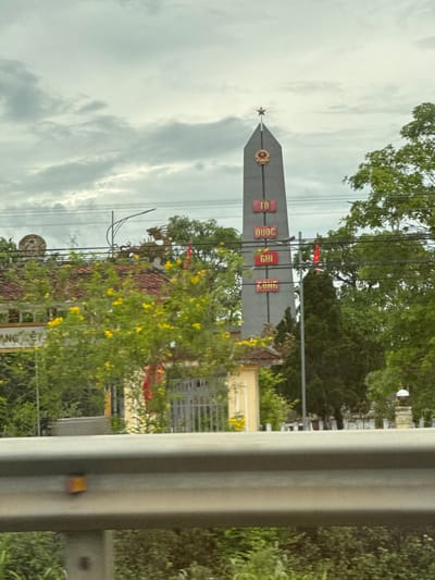 The image shows a monument in Hải Lăng Commune, Vietnam. The monument is a tall, gray obelisk topped with a star. The obelisk has inscriptions in Vietnamese that read "TỔ QUỐC GHI CÔNG" which translates to "The Nation Remembers Your Merits." A red flag with a yellow star, the flag of Vietnam, flies behind the monument. The monument is surrounded by trees and a cloudy sky. In the foreground, a portion of a metal guardrail is visible. The overall scene suggests a solemn memorial site.