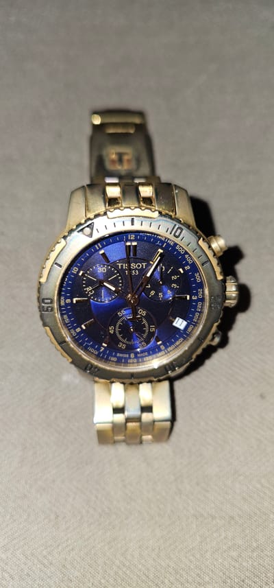 This is a close-up, top-down shot of a Tissot chronograph wristwatch with a blue dial. The watch features a stainless steel and gold-toned case and bracelet. The bezel is marked with tachymeter scales and has gold-toned numerals. The dial has three sub-dials for chronograph functions, a date window, and luminescent hour and minute hands. The brand name "TISSOT" is clearly visible on the dial. The watch is positioned on a textured, light-colored surface, possibly a fabric. There are shadows cast on the surface, suggesting indoor lighting. The image appears to be taken in Makati, Philippines, but there are no specific visual cues in the image to confirm this location, other than the provided context. The image captures a still object, a wristwatch, with no discernible activity or event taking place. The focus is on the details of the watch's design and craftsmanship.