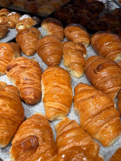 This is a close-up, eye-level shot of a large assortment of golden-brown croissants arranged on a light gray surface, likely a baking sheet or display tray. The croissants are various sizes and shapes, with many showing the characteristic layered pastry texture. Some appear to be plain, while others have a glaze that makes them glisten. In the background, other pastries can be seen, including some that are dusted with white sugar or coconut. The overall impression is that of a bakery display. The image does not provide enough information to determine the specific location in Holon, Israel.