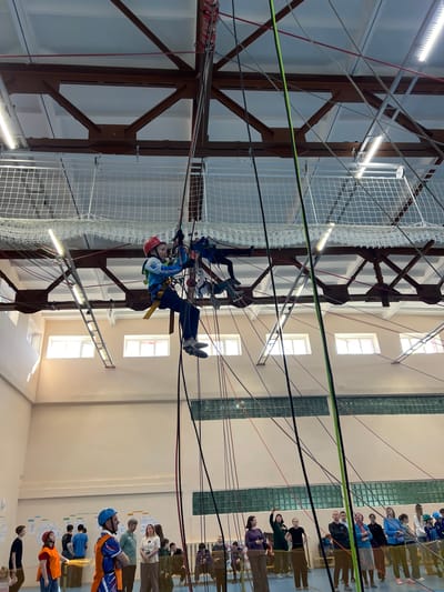 The image shows an indoor climbing structure in Berezniki, Russia. Two individuals are navigating a high-rope course. The person closer to the viewer wears a red helmet, a blue jacket with white lettering, and a yellow harness. The other person wears a blue helmet. They are suspended by ropes and cables connected to an overhead metal framework. A safety net surrounds the structure. In the background, a group of people are observing. The building's interior is lit by rows of fluorescent lights, with windows visible on the right wall.