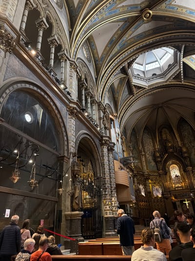 This media file captures the grand and ornate interior of a basilica or monastery, viewed from a low-angle perspective looking upwards. The scene is dominated by the high, vaulted ceilings, which are intricately decorated with elaborate gold and blue patterns on a light background. Majestic ribbed arches curve overhead, leading towards a central octagonal skylight or cupola that allows natural light to enter.
On the right side of the image, a large, complex pipe organ with numerous visible pipes is integrated into the upper architectural levels. Beyond the organ, the sanctuary or altar area is brightly lit and lavishly gilded, featuring religious artwork, statues, and a prominent cross. Ornate columns, balustrades, and decorative balconies line the upper sections of the interior.
In the foreground and lower right, a group of casually dressed people, likely tourists or visitors, are gathered, looking towards the front of the church or upward at the elaborate architecture. A red barrier rope is visible, delineating a path or restricted area for visitors. Decorative chandeliers and hanging lamps provide additional illumination throughout the interior.
The opulent architecture, with its detailed embellishments and prominent pipe organ, suggests a significant religious site, characteristic of locations like the Montserrat Monastery near Vacarisses, Spain. The scene appears to be taking place during the daytime, with a combination of natural light from the skylight and artificial lighting highlighting the interior's rich details. Small, unreadable red informational signs are visible among the crowd.